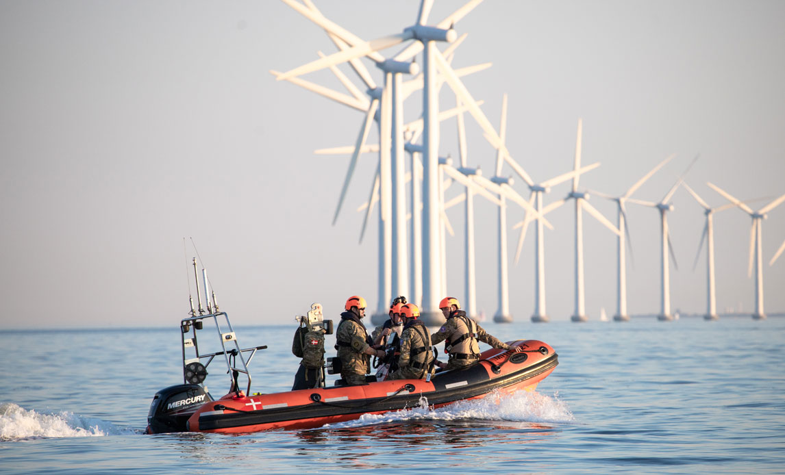 Åben hav. I baggrunden ses ca. 15 vindmøller. I forgrunden ses en rød og sort gummi-speedbåd med fire passagerer om bord.